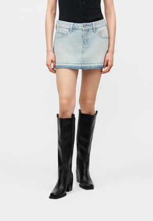 Femeie purtând o fustă mini din denim albastru deschis, un top negru fără mâneci și cizme negre din piele până la genunchi, stând în picioare pe un fundal simplu.