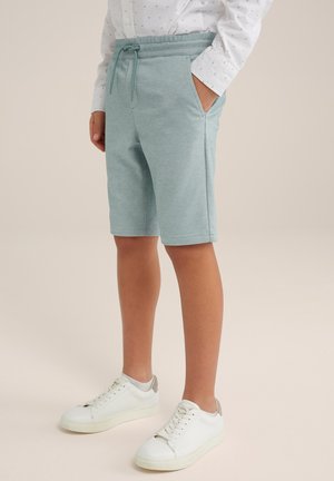 Enfant portant un short bleu clair à cordon, une chemise blanche à manches longues avec de petits motifs foncés, et des baskets blanches, debout les mains dans les poches.