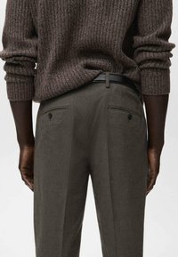 Persona de piel oscura que lleva un suéter marrón acanalado y pantalones grises entallados con bolsillos traseros abotonados, de pie con las manos a los lados.