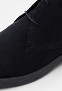 Chaussure en suedine marine avec un bout arrondi, une couture discrète et une semelle en caoutchouc texturé. Présente un détail de laçage minimal et un logo en relief.