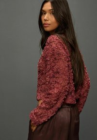 Burgundy floral-embroidered cropped top with layered petals and long sleeves, paired with brown silky trousers. Smooth texture, structured design.
