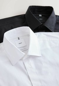 Deux chemises habillées soigneusement pliées, une blanche et une noire, toutes deux avec des cols pointus et des étiquettes de marque visibles portant la mention "NEXT."