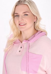 Světle růžová mikina s pruhovanou kapsou a stahovací kapucí. Obsahuje dva knoflíky na výstřihu a žebrovanou texturu.