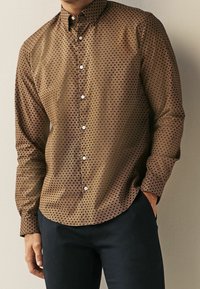Camisa marrón de botones con un patrón geométrico de círculos superpuestos, que presenta un cuello clásico, mangas largas y botones blancos.