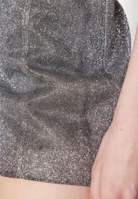 Tejido plateado metálico con una superficie texturada y brillante. Los bordes de la prenda son suaves, y una mano es visible cerca de la esquina inferior derecha.