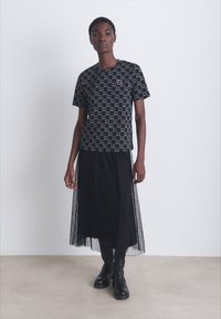 Μαύρη γραφική μπλούζα με επαναλαμβανόμενο σχέδιο λογοτύπου, συνδυασμένη με μια πλισέ μαύρη μίντι φούστα και μαύρες μπότες μέχρι το γόνατο, σε ουδέτερο φόντο.