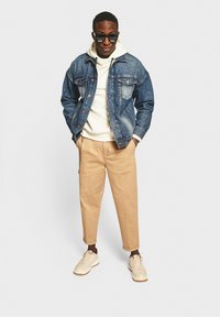 Denimjacket met blauwe tint over een witte hoodie, gecombineerd met beige broeken en lichte sneakers. Accessoires zijn onder andere donkere zonnebril.