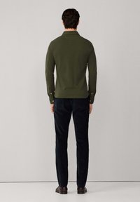 Camisa de manga larga verde oliva con cuello, corte ajustado y puños abotonados, combinada con pantalones oscuros de color marino y zapatos marrones.