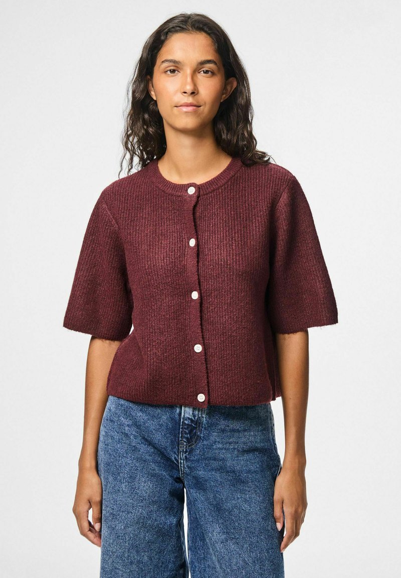 Bordeaux geribbelde cardigan met korte mouwen, voorzien van een ronde hals en crème knopen, gecombineerd met blauwe denim jeans.