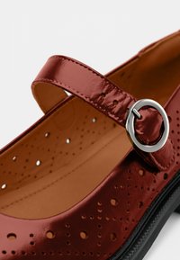 Κόκκινο παπούτσι Mary Jane με διάτρητα διακοσμητικά σχέδια, ρυθμιζόμενο λουράκι με στρογγυλή ασημένια αγκράφα, μπεζ εσωτερικό και μαύρη σόλα.