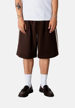 Person iført mørkebrune, vide shorts med hvide sidelister, hvide sokker, mørke læder loafers, en hvid ribbet skjorte og sølvsmykker på hænderne.