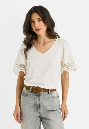 Mujer con cabello oscuro que lleva una blusa blanca con escote en V y mangas de encaje, vaqueros grises y un cinturón marrón, de pie frente a un fondo liso.