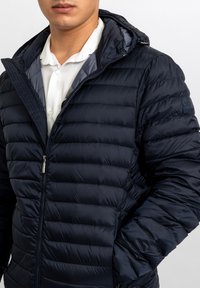 Giacca imbottita blu navy con design trapuntato, chiusura frontale con zip e cappuccio. Presenta un logo sulla spalla sinistra e una texture liscia e lucida.