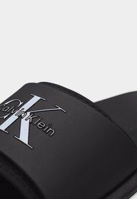 Μαύρη σαγιονάρα Calvin Klein slide με ανάγλυφο λευκό λογότυπο "CK" και κείμενο "Calvin Klein" στο λουράκι, κοντινή γωνιακή όψη.