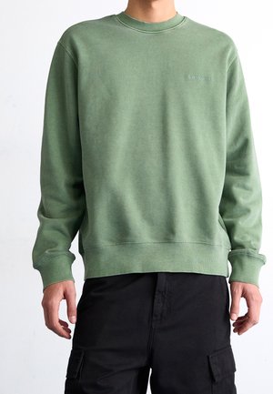 Person trägt einen grünen Carhartt-Pullover und schwarze Cargohosen und steht vor einem schlichten weißen Hintergrund.