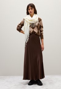 Donna che indossa un maglione marrone a rombi, una lunga gonna marrone scuro, una sciarpa frangiata bianca e scarpe nere, in piedi davanti a uno sfondo semplice.
