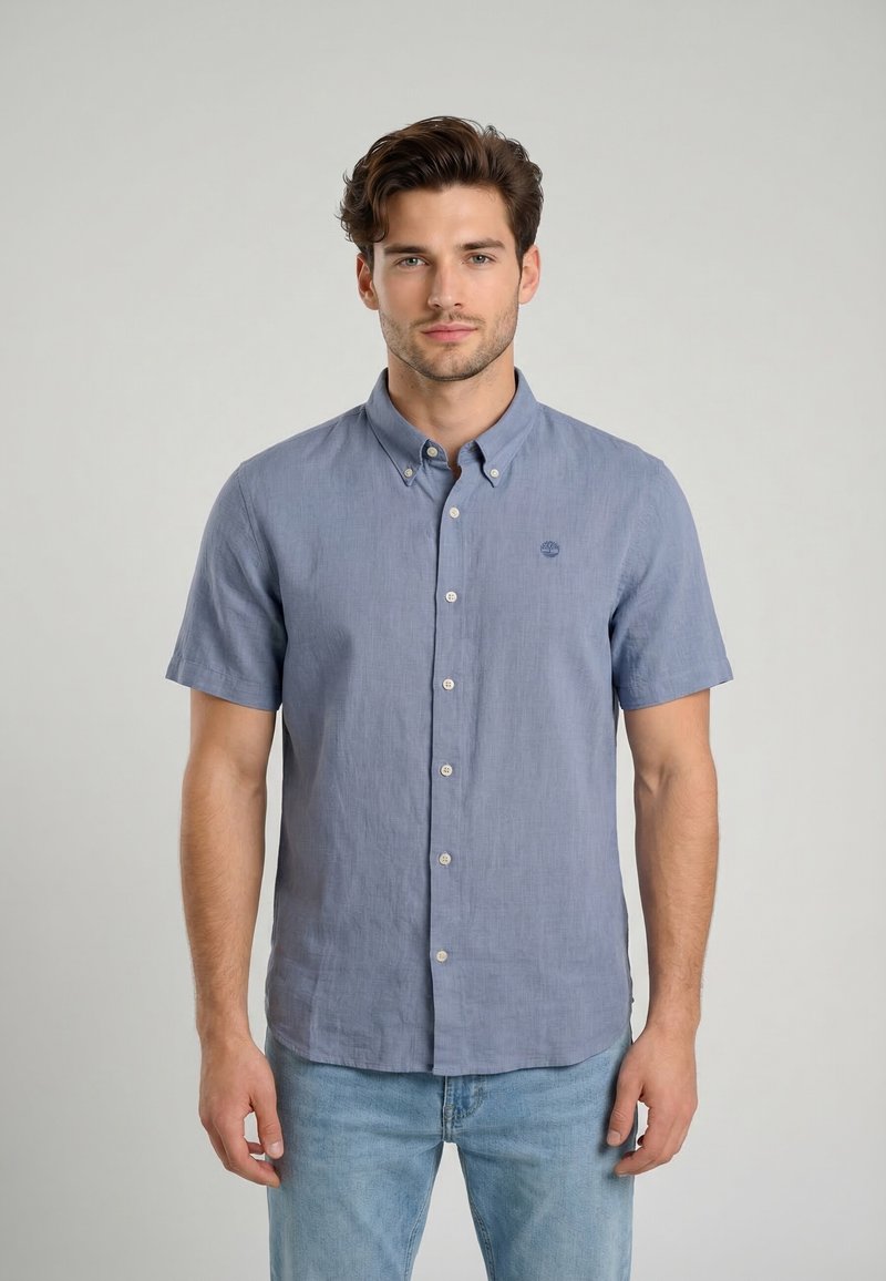 Homme portant une chemise bleue à manches courtes boutonnée et un jean bleu clair, debout face à l'avant sur un fond uni.