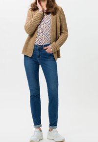 Pletený béžový kardigan přes vzorovanou blůzu v modré, hnědé a krémové, spojený s tmavě modrými džíny a bílými teniskami. Viditelné jednoduché designové detaily.