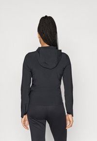 Melna viegla kapucēta jaka ar pieguļošu dizainu, garām piedurknēm un gludu tekstūru. Skats no aizmugures.