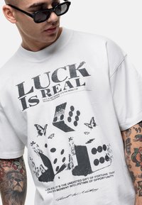 Witte katoenen t-shirt met een grafische print van de tekst "HEDEN IS ECHT," dobbelstenen, vlinders en ingewikkelde patronen in zwart. Relaxte pasvorm.