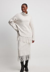 Helle beige Strickpullover mit hohem Kragen und fransigem Saum, kombiniert mit einem passenden fransigen Rock. Beide haben eine weiche, strukturierte Optik.