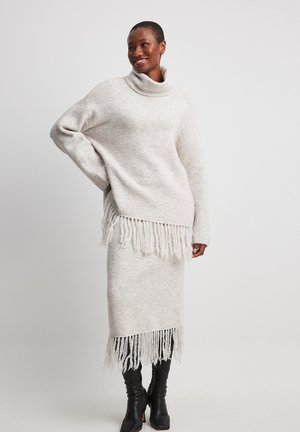 Lys beige strikket sweater med høj krave og frynsedetailer forneden, parret med en matchende frynset nederdel. Begge har et blødt, struktureret udseende.