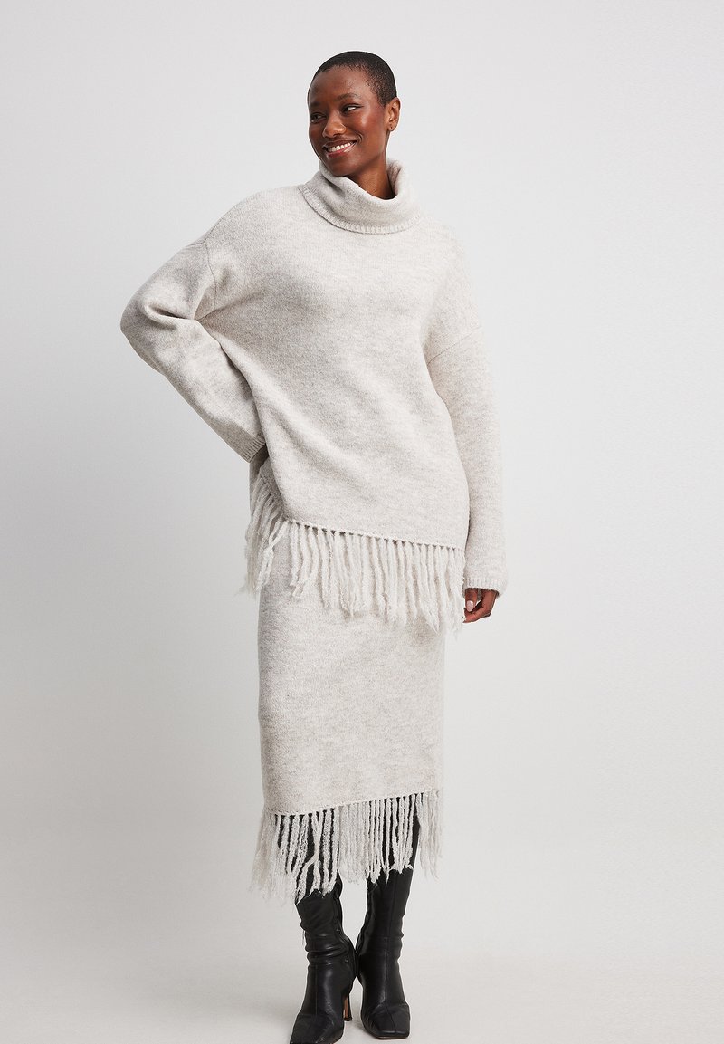Helle beige Strickpullover mit hohem Kragen und fransigem Saum, kombiniert mit einem passenden fransigen Rock. Beide haben eine weiche, strukturierte Optik.