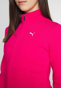 Femme portant une veste zippée rose vif avec le logo Puma blanc sur la poitrine, cheveux bruns raides, fond gris neutre.
