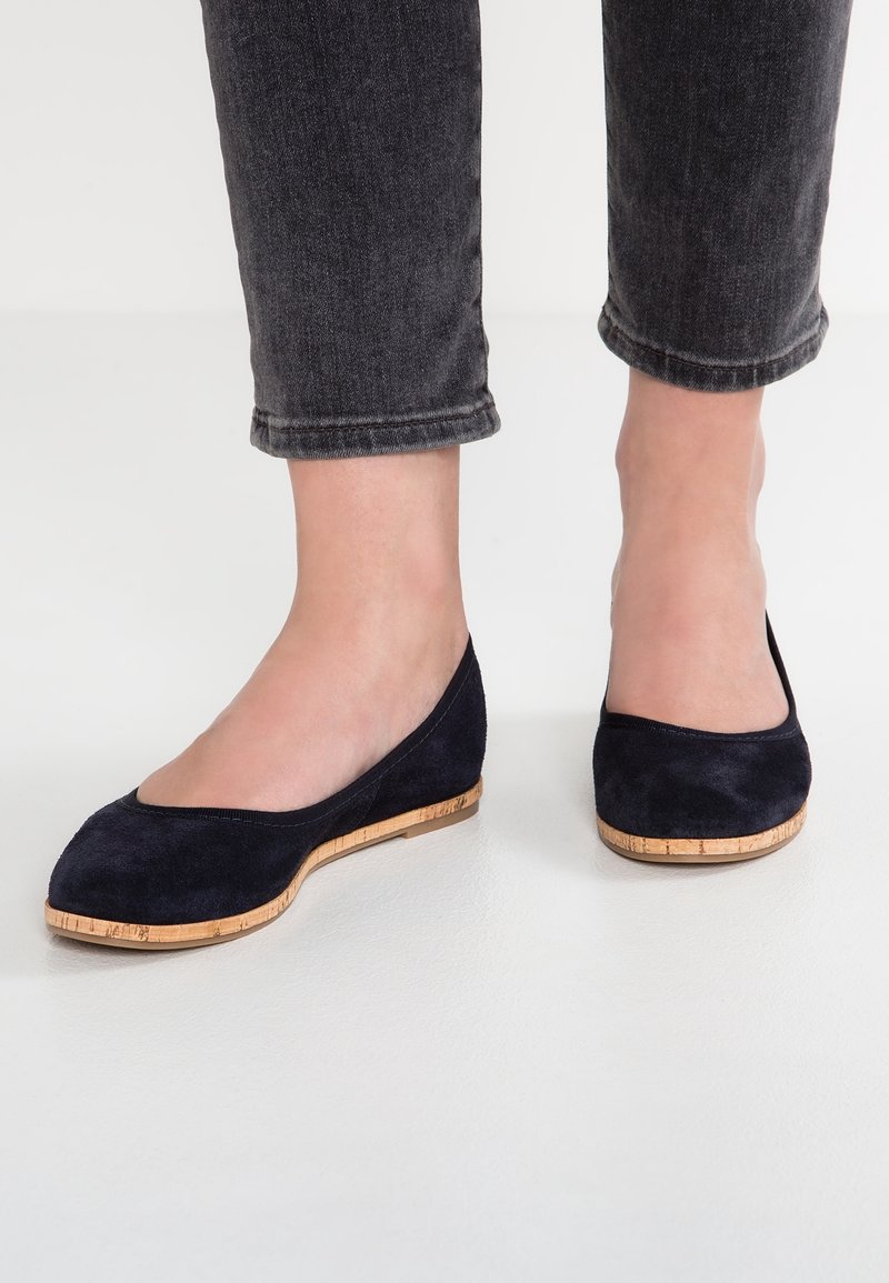 Personne portant des ballerines en daim noir avec des semelles en liège et un jean foncé retroussé, debout sur un sol blanc.