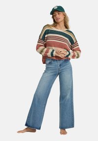 Maglione a righe in ruggine, crema e blu, abbinato a jeans in denim azzurro chiaro a gamba larga. Il modello indossa un cappellino verde.