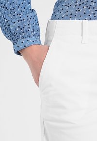 Mano nella tasca laterale di pantaloni bianchi, indossando una camicia a maniche lunghe blu con pois astratti in bianco e nero.