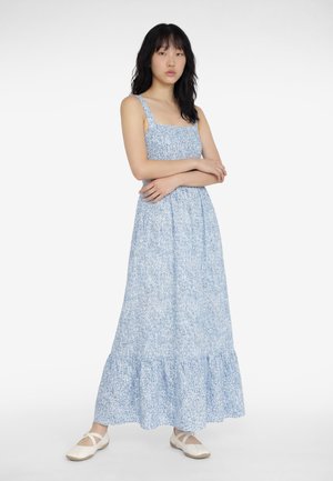 Kobieta o czarnych włosach, ubrana w bezrękawnikową, jasnoniebieską suknię maxi w kwiaty i białe baleriny, stojąca z założonymi rękami na białym tle.