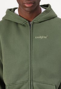 Grüne Zip-Hoodie aus weichem Material, mit einer frontalen Tasche und minimalistischer Markenkennzeichnung in passender Farbe. Akzentuiert mit einem silbernen Reißverschluss.