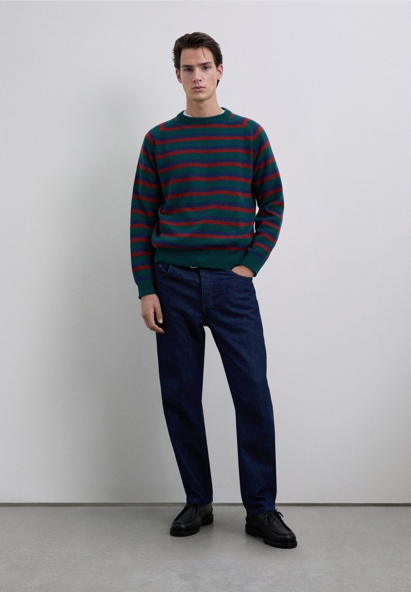 Groen en rood gestreepte trui met een ronde hals, gecombineerd met donkerblauwe spijkerbroek. Model draagt zwarte schoenen en staat tegen een lichte achtergrond.