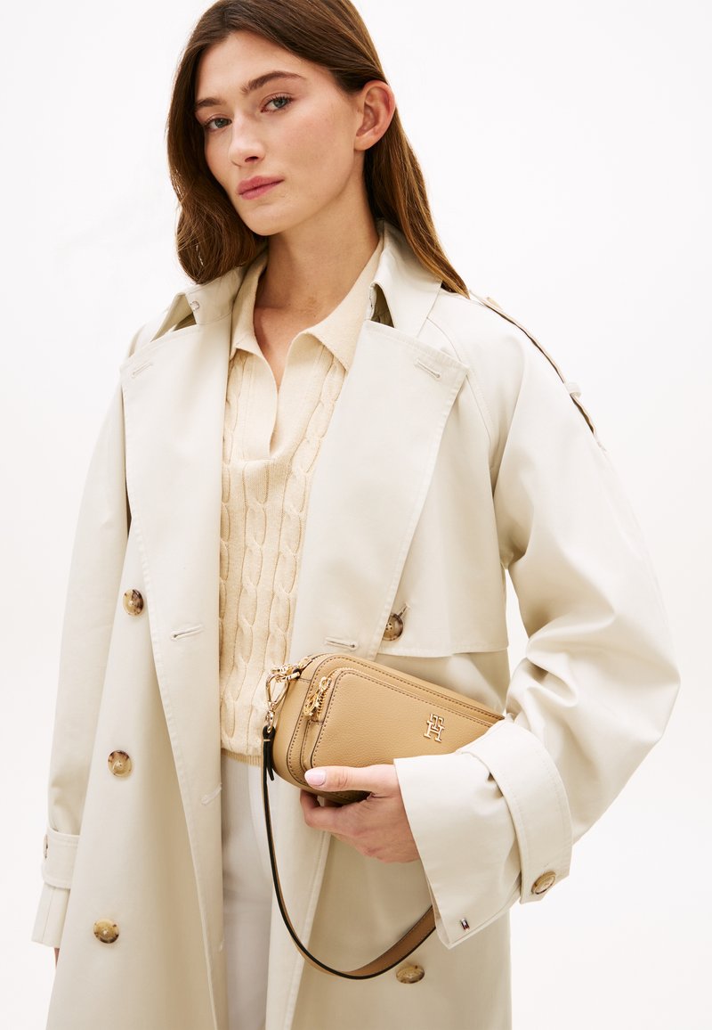 Γυναίκα με μακριά καστανά μαλλιά, που φοράει ένα μπεζ παλτό και πουλόβερ με πλέξη καλώδιο, κρατώντας μια μπεζ δερμάτινη τσάντα χιαστί με χρυσές λεπτομέρειες.