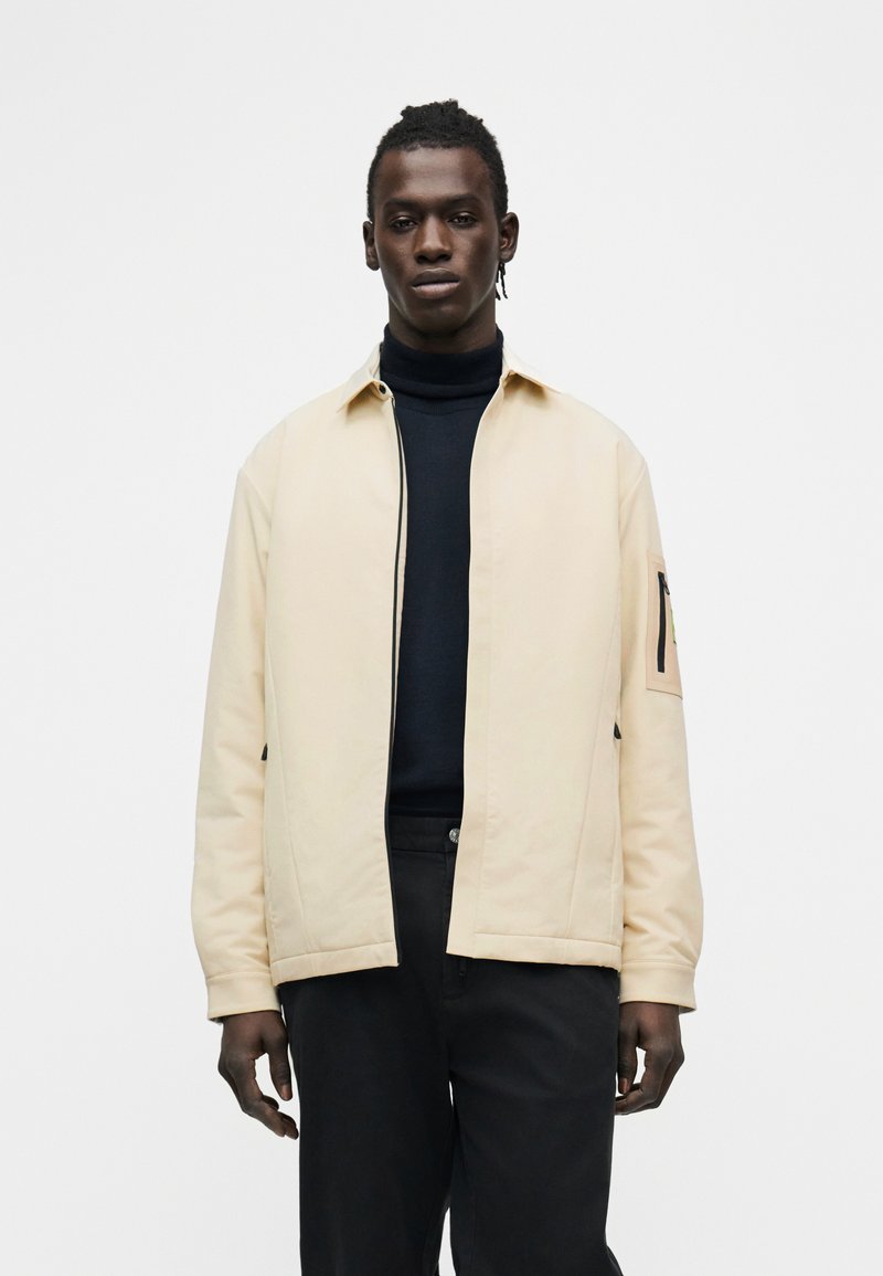 Homme portant une veste beige à fermeture éclair sur un col roulé noir et un pantalon noir, debout devant un fond blanc uni.