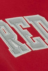 Roter Stoff mit einem auffälligen grauen gestickten Text, der ein strukturiertes, geschichtetes Design und kontrastierende Stickdetails aufweist.