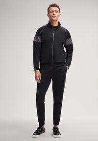 Schwarzer Sportanzug mit einer Reißverschlussjacke, die strukturierte Akzenten an den Schultern aufweist, kombiniert mit schmal zulaufenden Hosen und schwarzen Turnschuhen.