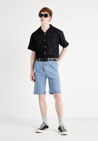 Camisetta nera a maniche corte con texture, pantaloni corti in denim azzurro, cintura nera, sneakers nere, calzini grigi, occhiali da sole oversize, sfondo minimalista.