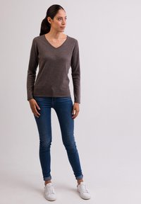 V-ringad, långärmad brun topp i mjukt material, kombinerad med blå slim fit jeans och vita sneakers. Modellen står mot en neutral bakgrund.