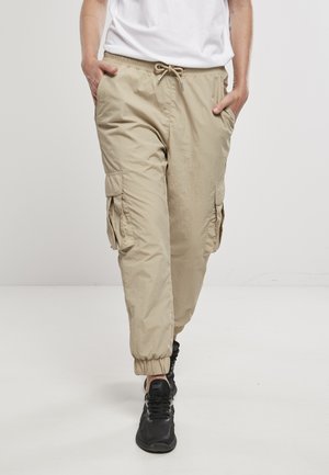 Person trägt beige Cargohose mit elastischen Bündchen und schwarzen Sneakers, Hände in den Taschen, geht vorwärts vor einfachem Hintergrund.
