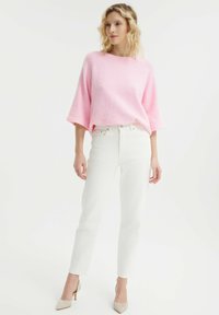 Maglione rosa, oversize, con texture e maniche larghe, abbinato a jeans bianchi slim-fit e scarpe con tacco beige.
