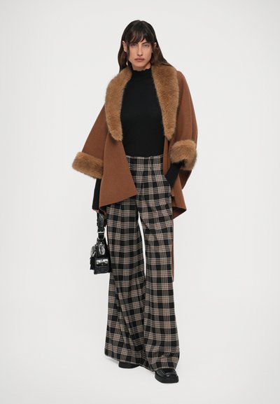 Mujer vestida con una capa marrón con borde de piel, un suéter negro de cuello alto, pantalones de cuadros anchos y zapatos negros, sosteniendo un pequeño bolso negro frente a un fondo blanco.