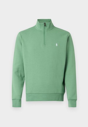 Homme portant un pull vert à fermeture éclair quart, chemise rayée en dessous, pantalon blanc et baskets blanches, debout les mains dans le dos.