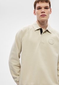 Bézs színű pulóver gallérral, fekete gombsorral és dombornyomott logóval. Texturált anyagból készült, raglán ujjal.