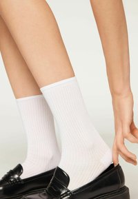 White ribbed socks made of cotton blend, featuring a knit design and a cuffed top, worn with black loafers. Smooth texture and fitted shape.