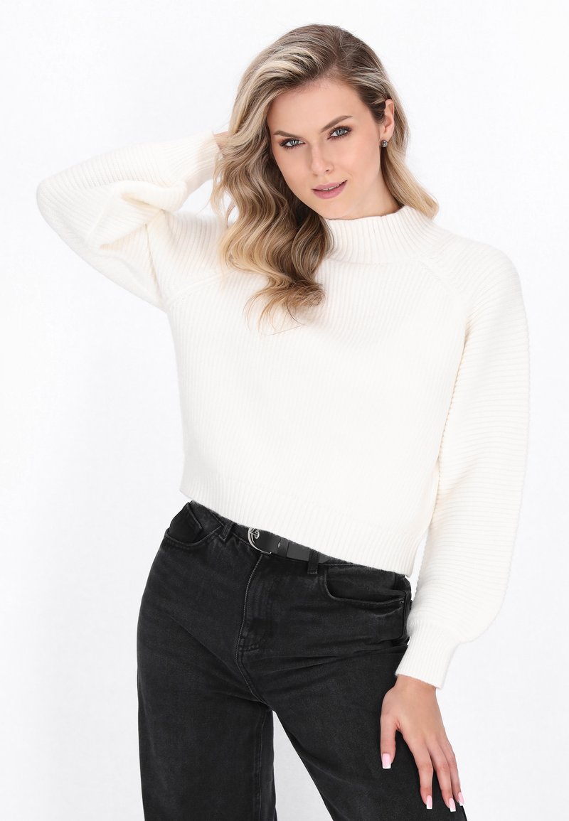 Hvid ribstrikket cropped sweater med høj hals og pufærmer, parret med sorte jeans og et sort bælte. Glat tekstur.