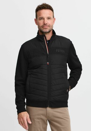 Black insulated jacket with a zip front, stand-up collar, and two side pockets. Features quilted design and subtle logo on chest.