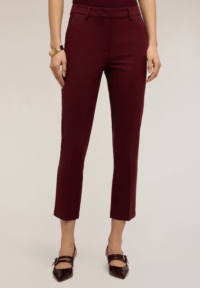 Vrouw draagt aansluitende bordeauxrode broek tot op de enkel, bijpassende puntige schoenen met bandjes en een gouden armband aan de linkerpols.