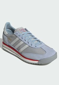 Light blue athletic shoe with suede and mesh materials, featuring three white stripes, a textured grey toe, and a red accent on the sole.